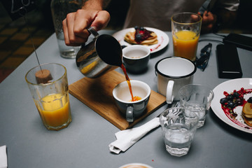 Delicious and healthy brunch or breakfast at fancy city hidden spot cafe, orange juice and wholewheat pancakes. Man pours organic filter specialty coffee from jar to cup. Food blogger