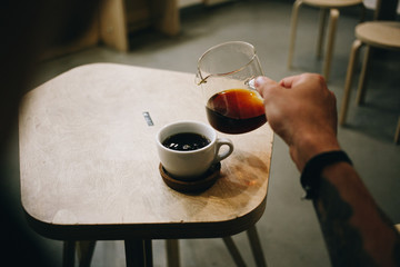 Cinematic vintage photo behind shoulder of man with arm tattoos pour filter coffee from glass jar into cup at specialty coffee shop or cafe. fancy caffeine drink addiction and millennial trend