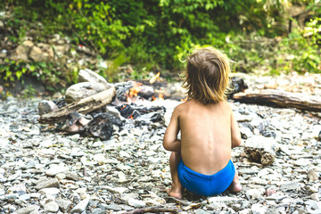 cute little boy in the forest sitting near the fire in the summer, view from the back