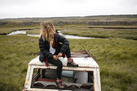 Young female explorer,traveller in puffy jacket,hiking boots and long blonde hair sits on top of old car during exploring of icelandic landscape.Cool hipster photo of millennial and social media trend