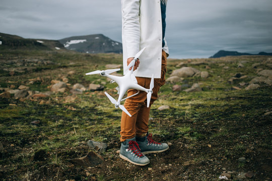 Close up of man in white hipster raincoat, tourist or social media influencer and blogger stand on rocky surface in iceland or norway during epic roadtrip, holds big professional drone