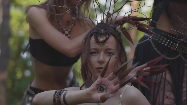 Portrait of young attractive woman in theatrical costume, hairstyle and make up of dryad dancing with painted eyes on palms in the forest showing performance or making ancient ritual