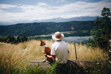 Young male hiker or adventurer tourist takes rest at scenic overlook at field on top of mountain together with pet dog basenji brown puppy. Healthy lifestyle and outdoor activities