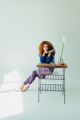 stylish redhead girl posing at table with calla flower and vases on grey