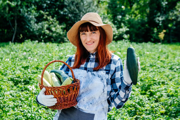 Portrait beautiful female farmer holds a zucchini in a wicker basket wearing a straw hat and surrounded by the many plants in her vegetable garden