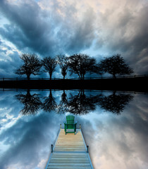 Mirror imaged of five trees along a roadway silhouetted against stormy clouds with an Adirondack chair sitting on a dock, Stowe Vermont, USA