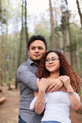 Fototapeta premium man hugging behind his girlfriend in the forest wearing casual clothes - hispanic couple