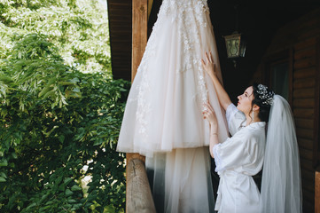 Beautiful bride brunette straightens white wedding dress, which hangs on a hanger. Morning, fees, wedding portrait of a cute girl. Photography and concept.