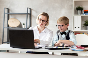 Young schoolboy in sweater and his charming teacher in white blouse sit behind desk and working with laptop