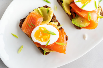 Healthy open sandwiches on multigrain wholegrain  toast with avocado, salmon, eggs, herbs, sunflower seeds over white plate on concrete background with copy space. Healthy protein food.