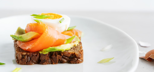Healthy open sandwiches on multigrain wholegrain  toast with avocado, salmon, eggs, herbs, sunflower seeds over white plate on concrete background with copy space. Healthy protein food. Wide banner.