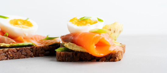Healthy open sandwiches on multigrain wholegrain  toast with avocado, salmon, eggs, herbs, sunflower seeds over white plate on concrete background with copy space. Healthy protein food. Wide banner.