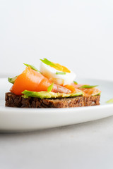 Healthy open sandwiches on multigrain wholegrain  toast with avocado, salmon, eggs, herbs, sunflower seeds over white plate on concrete background with copy space. Healthy protein food.