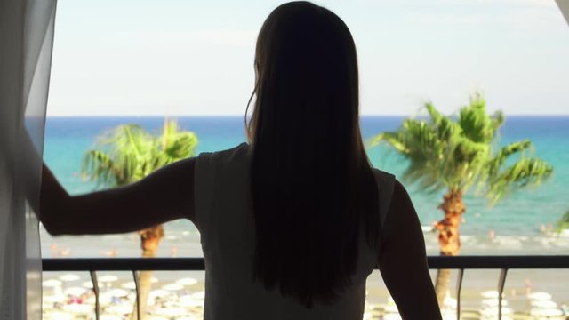 Young woman going out onto balcony and enjoying view from terrace. Female on vacation opening curtains and looking at Mediterranean sea. Wind swaying leaves on palm trees