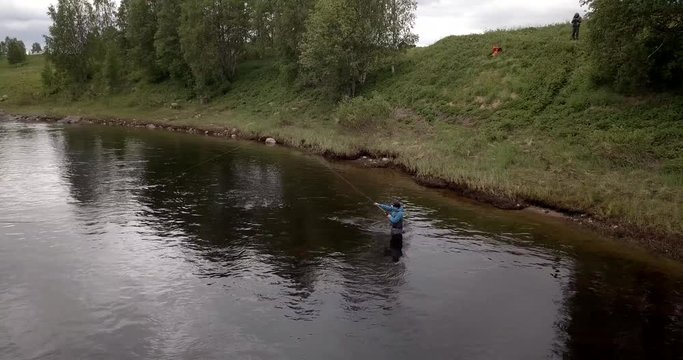 Aerial, drone shot, panning around a man fly fishing, in shallow water, close to the shore of the Tornionjoki river, on a cloudy day, in Pello, Meri-Lappi, Lapland, Finland