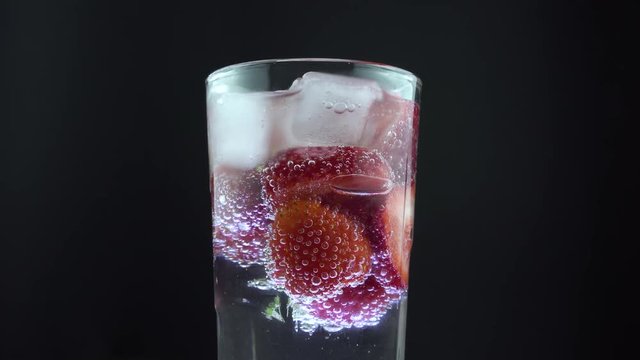 Tall glass soda water with ice cubes and strawberries, centre screen. Fizzy bubbles rising. Rotating in slow motion.