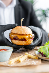 Young man in business suit and black gloves eating delicious juicy cheeseburger with beef chop. Burgers in the cafe with salad and fries, fast food.