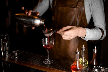Professional bartender pouring a red alcoholic drink from the steel shaker through the sieve