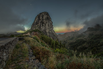 Naklejka premium Amazing sunrise near Los roques and famous Agando rock near Garajonay Park at La Gomera. Half moon on a background of gray and purple clouds. Relic laurels and heather on steep slopes. Canary, Spain