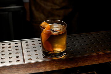 Golden cocktail in the glass with one stamped big ice cube and orange zest