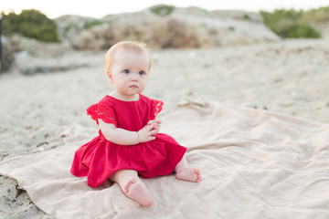 Cute baby girl in a red dress sitting and playing on the beach with sand