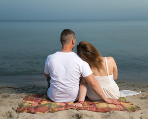 Back view of young happy couple,  sitting on seashore at sunset.