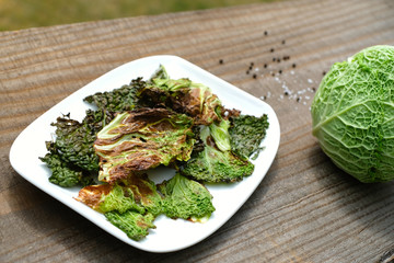 Close up selective focus high angle shot homemade tasty crispy savoy cabbage chips on a white plate on a vintage rustic table. Oven-baked leafy greens healthy leaves with salt, peppers and olive oil.