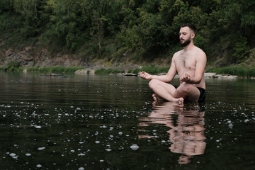 A guy in swimming trunks sits on the surface of the water in a river or lake. The concept of yoga, relaxation in the mountains, unity with nature, travel and lifestyle. Copy space.