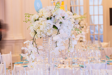Glass vases with long branches of white orchids and roses stand on dinner tables