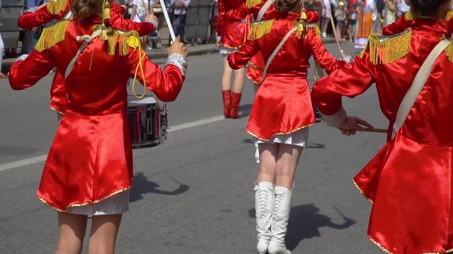 Young drummers line up and beat the melody. Street performance of festive march of drummers girls in red costumes on city street. Back view