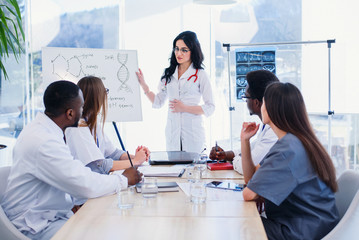 Beautiful female doctor wearing white coat and eyeglasses standing by flipchart and giving presentation to group of healthcare specialists. Medical team having a meeting in conference room in hospital