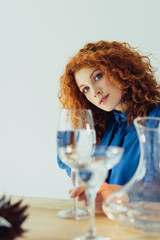 attractive stylish redhead woman posing near table with glasses of water isolated on grey