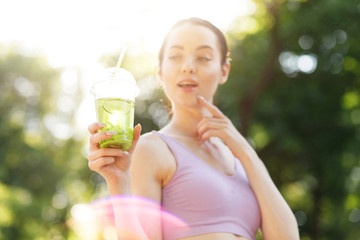 Young beautiful girl drinking green beverage with lime and kiwi from plastic cup take-out food. Smiling slim brunette woman with lemonade in park. Summer cold drinks.
