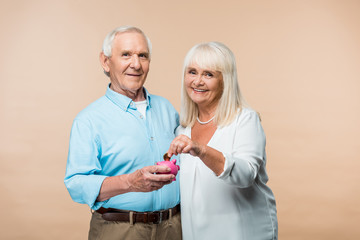 cheerful retired man holding pink piggy bank while senior wife putting coin isolated on beige