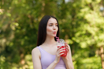 Young beautiful girl drinking fresh juice from plastic cup take-out food. Smiling slim brunette woman with berry lemonade in park. Summer cold drinks.