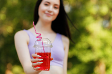 Young beautiful girl drinking fresh juice from plastic cup take-out food. Smiling slim brunette woman with berry lemonade in park. Summer cold drinks.