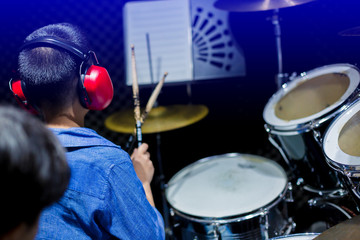 Asian boy put blue shirt and red headphone to learning and play drum set with wooden drumsticks in music room. Teacher teach boy to pay drum set. The concept of musical instrument, Selective focus.