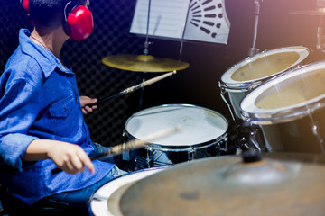 Fototapeta na wymiar Asian boy put blue shirt and red headphone to learning and play drum set with wooden drumsticks in music room. The concept of musical instrument, Side view to drum player and selective focus.