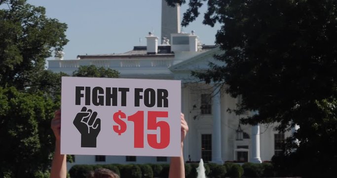 A man holds a Fight for $15 minimum wage protest sign in front of the White House on a sunny summer day.  	