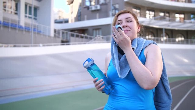 Close-up portrait of body positive happy overweight female runner walking along stadium track after morning workout. Joyful fat woman with towel and bottle relaxing satisfied with good training