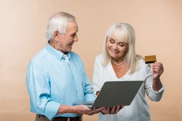 cheerful retired woman holding credit card near senior husband with laptop isolated on beige