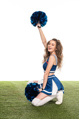 smiling cheerleader girl in blue uniform sitting with pompoms on green field isolated on white