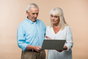 retired woman gesturing while looking at laptop near senior husband isolated on beige