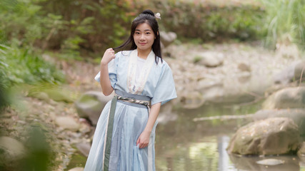 Beautiful Asian woman in cyan Chinese costume clothes hanfu beside of stream and looking at camera in peaceful smile, traditional ancient Chinese beauty.