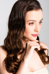 Naklejka premium beautiful brunette woman with curls and makeup looking away with hand near chin isolated on grey