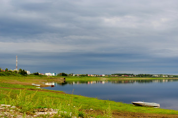 A small boat stands on the shore on the Northern Yakut river vilyu near the village of ulus Suntar with houses and a communication tower along the water under a cloudy sky.