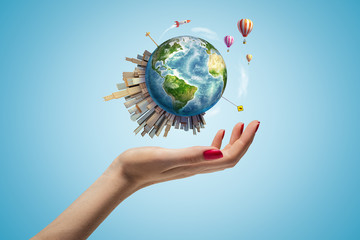 Woman's hand levitating small Earth with big modern city popping up on one side and hot-air balloons and rocket in air on light-blue background.