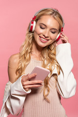 beautiful blonde happy woman with headphones using smartphone isolated on pink