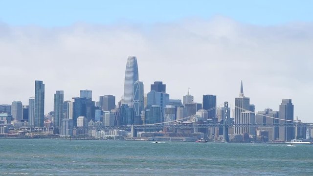 HD video zooming in on downtown San Francisco and the bay bridge in the summer, heavy marine layer wind blowing clouds through the city reflecting in windows of the buildings as ships sit in the bay. 