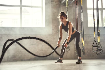 Photo sur Plexiglas Fitness Préparation au concours. Jeune femme athlétique avec un corps parfait faisant des exercices de crossfit avec une corde dans la salle de gym.